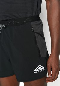 Shorts de trail negros de Nike con un panel lateral texturizado gris, cinturilla con cordón y un bolsillo con cremallera. Presenta un detalle de logo blanco.
