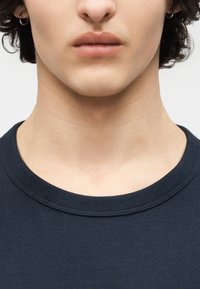 Gros plan sur le bas du visage, le cou et le haut de la poitrine d'une personne portant un t-shirt à col rond foncé, avec de petites boucles d'oreilles visibles.