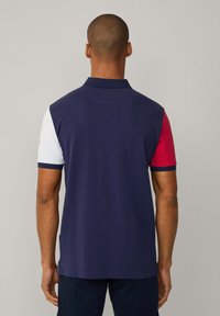Polo de color azul marino con mangas cortas; manga izquierda blanca y manga derecha roja. Cuenta con un cuello clásico y un dobladillo recto. Material de mezcla de algodón.