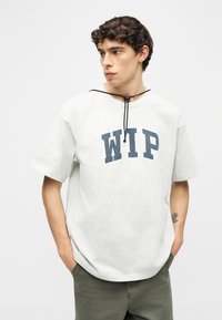 Νέος άνδρας με σγουρά μαλλιά που φοράει λευκό κοντομάνικο πουκάμισο με τα γράμματα "WIP", πράσινο παντελόνι και μαύρο κορδόνι στο λαιμό, με τα χέρια στις τσέπες.