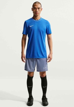 Hombre de pie vistiendo camiseta deportiva azul de Nike, pantalones cortos grises, calcetas negras hasta la rodilla y botas negras sobre un fondo claro y liso.