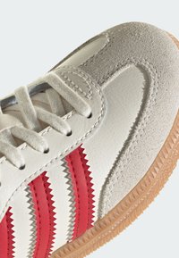 Witte sportschoen met rode gegroefde strepen, bovenwerk van leer en suede, textuur van rubberen zool en vetersluiting.