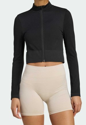 Vrouw die een zwart, langärmlig cropped jack met ritssluiting draagt en beige, hooggetailleerde, aansluitende biker shorts, tegen een effen achtergrond.