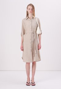Robe chemise en lin beige avec un devant à boutons, une taille cintrée et des manches retroussées. Accompagnée d'un petit sac à main blanc et de tongs.