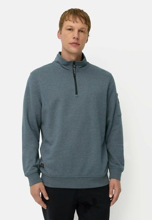Man draagt een blauw-grijze trui met een kwartritssluiting, lange mouwen en een zwarte broek, staand tegen een effen witte achtergrond.