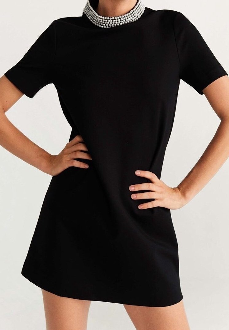 Femme portant une robe noire courte à manches courtes et un collier de perles multi-brins, mains placées sur les hanches.