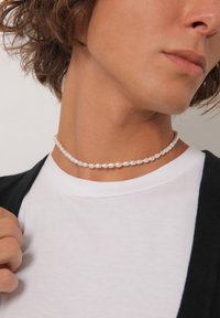 Choker tipo kaklo papuošalas, pagamintas iš mažų baltų perlų, su ovalia forma ir lygiu paviršiumi, uždėtas aplink kaklą ant baltos marškinėlių.