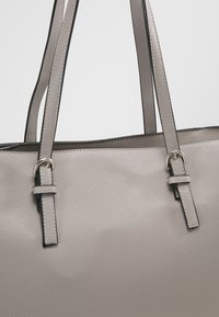 Sac à main gris clair texturé avec deux bretelles d'épaule réglables comportant des boucles en métal argenté sur un fond blanc uni.