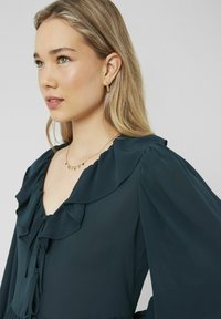 Blouse teal à manches longues avec un col volé, un détail à nouer et une texture de tissu douce. Un simple collier en or et des boucles d'oreilles créoles complètent le look.