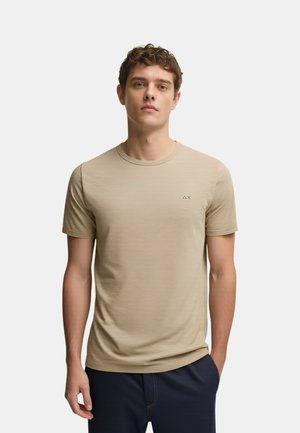 Giovane uomo con capelli ricci corti che indossa una t-shirt beige a maniche corte con scollo rotondo e pantaloni scuri, in piedi davanti a uno sfondo chiaro e uniforme.