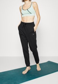Sujetador deportivo verde claro con detalles negros, combinado con pantalones deportivos negros que cuentan con bolsillos y un pequeño logo de Puma. Colocado sobre un tapete azul verdoso.