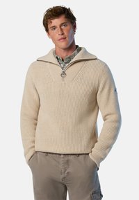Beige Strickpullover mit hohem Kragen und halber Reißverschluss, ausgestattet mit Rippstruktur und einem Logopatch am Ärmel.
