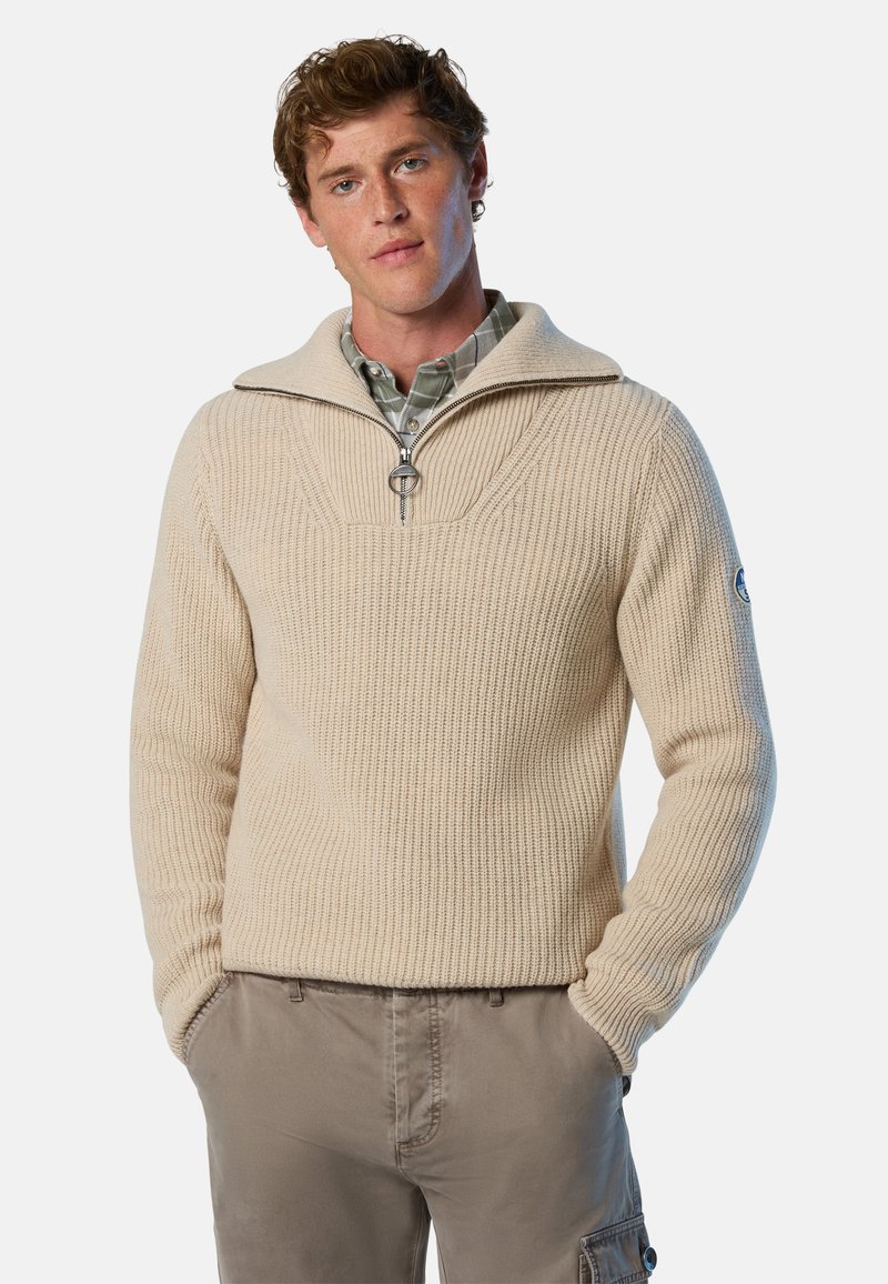 Beige Strickpullover mit hohem Kragen und halber Reißverschluss, ausgestattet mit Rippstruktur und einem Logopatch am Ärmel.