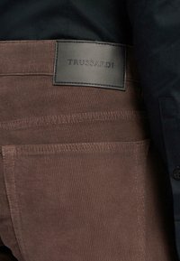 Pantalones de pana marrones con canalones verticales, que presentan una etiqueta de marca de cuero negro en la cinturilla trasera. Incluyen dos bolsillos traseros.
