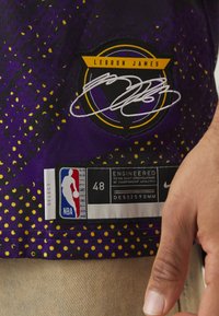 Canotta da basket viola con una toppa di LeBron James in giallo e nero, design a pois, etichetta NBA e etichetta della taglia. La texture appare liscia.