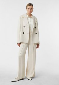 Cream double-breasted coat made of wool, paired with a light-colored shirt and wide-leg trousers. Flat-toed shoes complete the outfit.