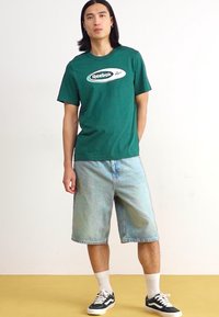 Žalias medvilninis marškinėliai su Reebok logotipu, derinami su nublukusiais džinsiniais šortais, baltais kojiniais ir juodais sportbačiais. Modelis stovi prieš paprastą foną.