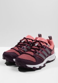 Zapatillas deportivas moradas y rosas con parte superior de malla, detalles texturizados, suela de goma y tres franjas grises en el lateral.