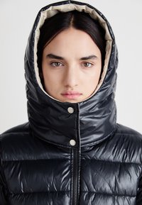 Μαύρο μπουφάν πούφας με ψηλό, επενδεδυμένο καπέλο με κρέμα. Διαθέτει κούμπωμα με snaps και φερμουάρ μπροστά, αναδεικνύοντας μια καπιτονέ υφή.