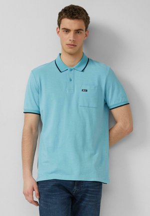Jonge man die tegen een muur staat, gekleed in een lichtblauw poloshirt met zwarte bies en donkerblauwe spijkerbroek, linkerhand in de zak.