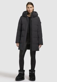 Piumino nero con cappuccio, con design trapuntato, zip frontale e bottoni a pressione. Indossato sopra leggings neri lucidi e stivali.