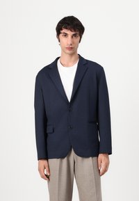 Marineblå blazer laget av teksturert stoff, med to knapper, en notched lapel og sidelommer, kombinert med lyse bukser.