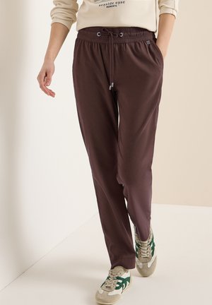 Persona che indossa pantaloni marrone scuro con coulisse, felpa beige con scritta e sneakers beige con dettagli verdi che cammina all'interno.