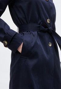 Τζιν μαντό σε ναυτικό μπλε χρώμα με μεγάλα κουμπιά, ζώνη στη μέση που δένει, κουμπιά στα μανίκια και ένα χέρι τοποθετημένο στην πλαϊνή τσέπη.