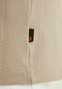 Pull tricoté beige avec un motif en zigzag texturé. Présente une étiquette noire avec un accent carré marron. Détail de couture visible le long du bord.