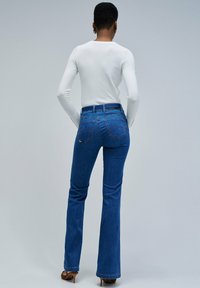 Femme se tenant de dos, portant un haut blanc à manches longues, un jean flare taille haute bleu et des sandales à talons marron contre un fond uni.