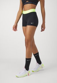 Zwarte Nike Pro shorts met een neongele tailleband, voorzien van het Nike logo. Gepaard met witte schoenen en zwarte sokken, wat een slanke atletische uitstraling geeft.