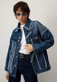 Persona, kas valkā nolietotu dūrienu denim jaku, baltu kreklu, tumšas bikses un taisnstūrveida saulesbrilles, pozē pret vienkāršu fona.