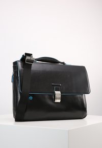 Borsa a tracolla in pelle nera con cuciture blu, tracolla regolabile, chiusura con gancio metallico e tasca laterale con zip. Texture liscia.
