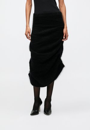 Gonna nera e morbida con design drappeggiato, caratterizzata da una texture soffice e un orlo asimmetrico. Indossata con tacchi alti neri e collant.