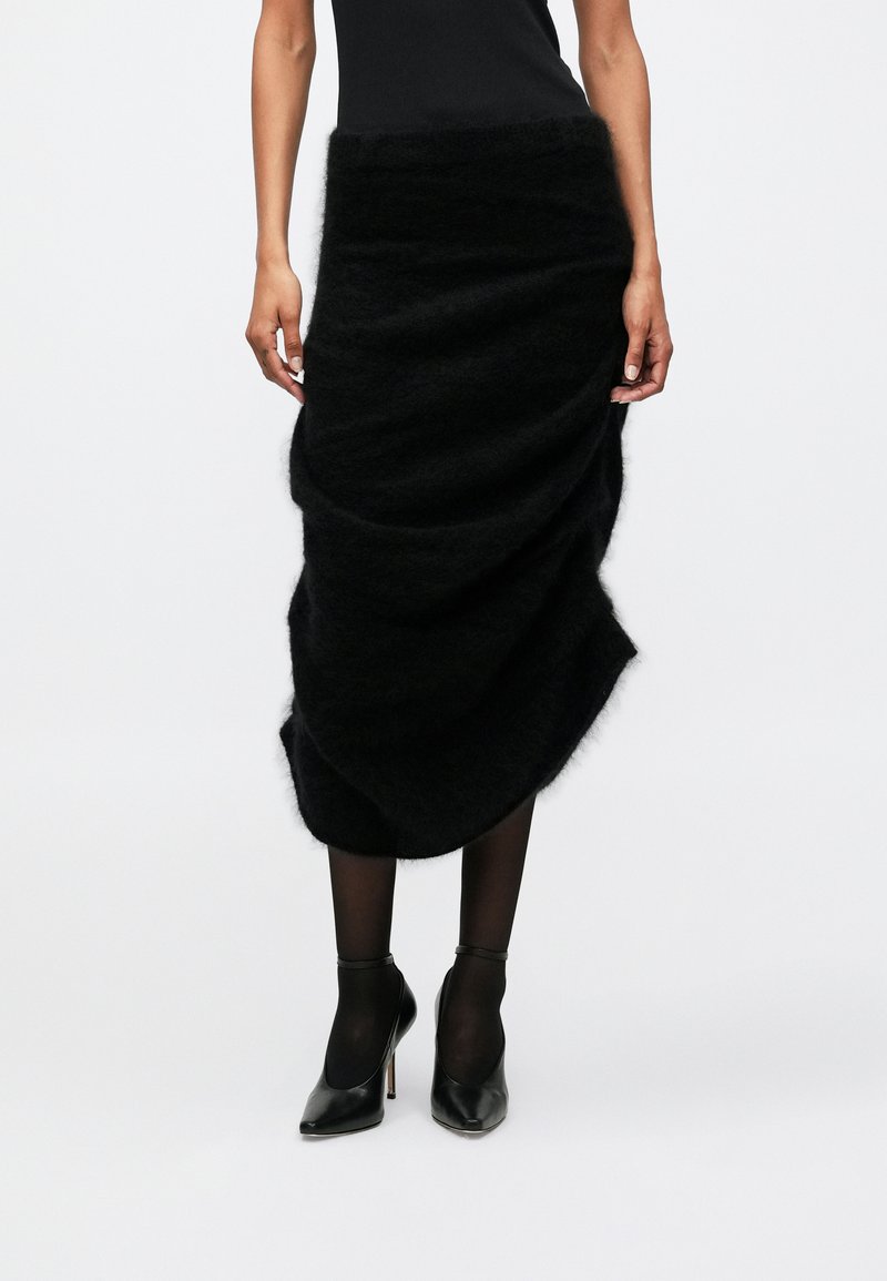 Saia preta, fofa, com um design drapeado, apresentando uma textura suave e uma bainha assimétrica. Usada com saltos altos pretos e collants.