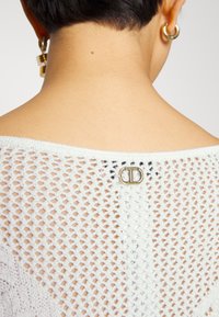 Haut en tricot blanc avec un design à maille ouverte, une fermeture à boucle visible à l'arrière et un col lisse. Détail de tissu texturé évident.