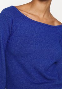 Modrý pletený svetr s výstřihem přes ramena a žebrovanou strukturou. Má dlouhé rukávy a měkkou, lehkou látku.