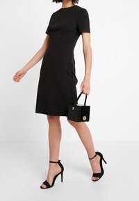 Schwarz enganliegendes Kleid mit kurzen Ärmeln und knielangem Saum. Kombiniert mit schwarzen Riemchenheels und einer kleinen glänzenden schwarzen Handtasche mit einem goldenen Akzent.