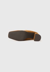 Zapato de tacón alto de ante con punta cuadrada, parte superior marrón y suela de goma negra texturizada. El tacón es ancho y robusto para mayor estabilidad.