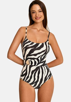 Femme souriante, portant un maillot de bain une pièce à rayures zébrées noir et blanc avec des bretelles fines et une boucle de ceinture décorative à la taille.