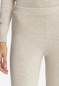 Leggings beige fabriqués dans un matériau doux et texturé. Dotés d'une taille sans couture et d'une finition lisse. La main est placée près du tissu.