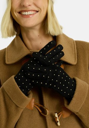 Gants noirs avec des clous dorés, fabriqués en matériau doux, portés avec un manteau en laine marron avec des fermetures à toggle. Les gants sont de longueur doigts, style ajusté.