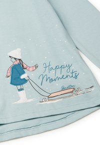 Dívka v zimním oblečení táhnoucí sáňky ve sněhu s textem "Šťastné okamžiky" na světle modré látce.