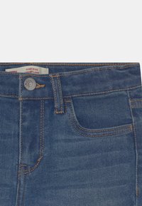 Vaqueros de mezclilla azul de talle alto con corte acampanado. Presentan un solo bolsillo y costuras en contraste de color amarillo. Cierre con botón plateado con detalle de logotipo.