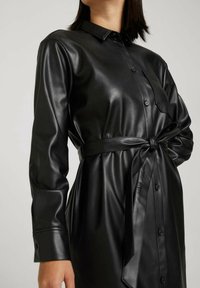 Schwarzes Hemdkleid aus Kunstleder mit durchgehender Knopfleiste, langen Ärmeln und einem gebundenen Taillengürtel, das eine glatte Textur und ein glänzendes Finish aufweist.
