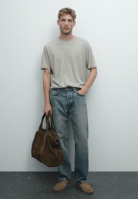 Man wearing light gray T-shirt, blue jeans, and brown shoes, holding a large brown suede handbag, standing against a plain wall.