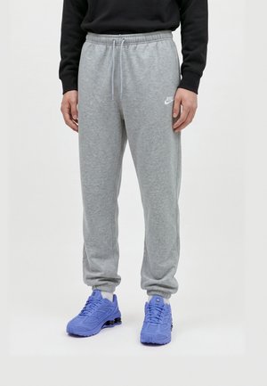 Le mannequin porte un pantalon de jogging Nike gris avec un cordon de serrage à la taille et des bas resserrés, associé à un haut noir et à des baskets bleues.