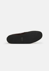 Zapato marrón oscuro con una suela de goma negra texturizada, que presenta un patrón de diamantes, y un material superior liso y ligeramente brillante.
