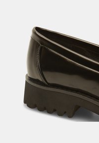 Scarpa in vernice nera con tacco basso e suola in gomma sagomata. Il design presenta una parte superiore liscia e una punta arrotondata.