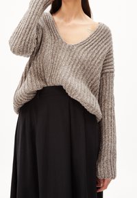 Pull beige en maille avec un col en V large et profond, une texture côtelée, et des manches longues, associé à une jupe noire taille haute.